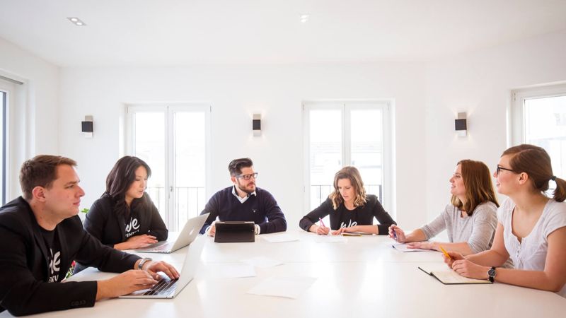 Группа мужчин и женщин, с ноутбуками, за большим столом разговаривают.