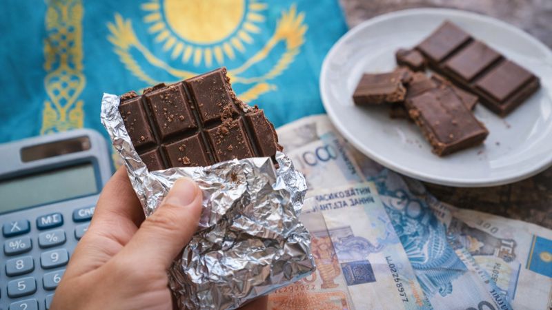 Шоколад, снижение потребления в 2025 году, руки держат шоколадную плитку на фоне тенге и флага Казахстана