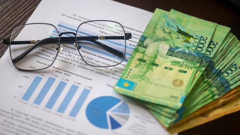 Купюры тенге на листах с диаграммами, рядом очки, аналитика о нацвалюте